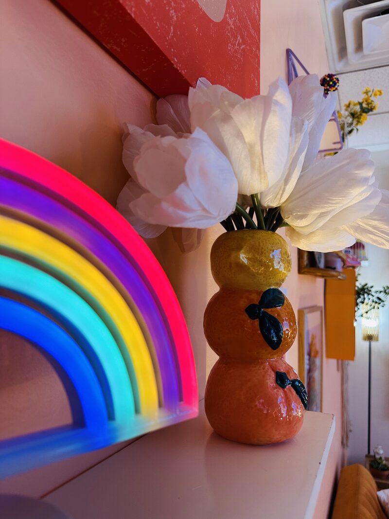 A neon rainbow beside a citrus-shaped vase of white flowers