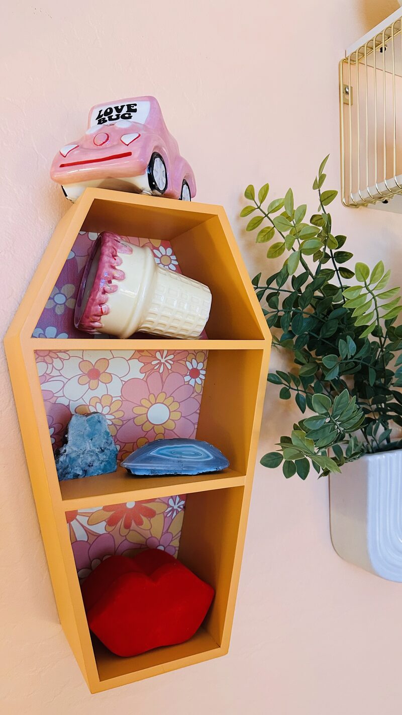 A coffin-shaped shelf with crystals, a Love Bug figurine, and a red lips cushion